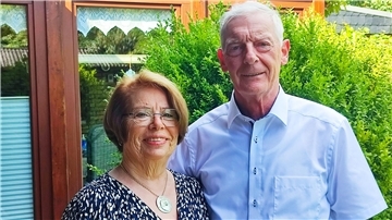 Eine Frau und ein Mann stehen in einem Garten vor einer Hecke. 
