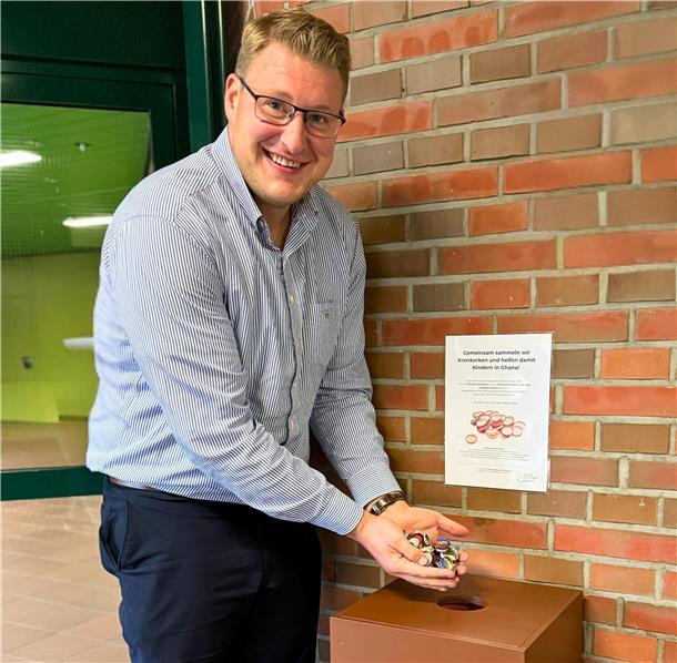 Ein Mann steht lächelnd neben einer braunen Sammelbox im Eingangsbereich eines Gebäudes und hält eine Handvoll Kronkorken. An der Wand hinter ihm hängt ein Hinweisplakat zur Sammelaktion für Kinder in Ghana.