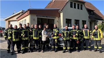 Gruppenfoto vor dem Gebäude der Freiwilligen Feuerwehr: Eine Gruppe von Feuerwehrleuten in Einsatzkleidung steht gemeinsam mit Bürgermeisterin Gabi Kasten, die eine Wärmebildkamera überreicht. Im Hintergrund sind Feuerwehrfahrzeuge und das Feuerwehrhaus zu sehen.