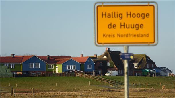 Hochsaison für den Halligkaufmann von Hooge Bunte Häuser sind auf einer Warft auf Hallig Hooge hinter dem Ortsschild zu sehen.