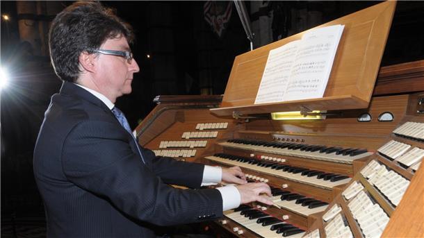 Ein Mann im Anzug spielt an einer großen Orgel mit mehreren Manualen und zahlreichen Registerzügen. Vor ihm liegt ein aufgeschlagenes Notenheft.