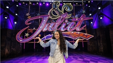 Chiara Fuhrmann steht auf einer Bühne vor einem Neon-Zeichen des Schriftzugs "& Julia"