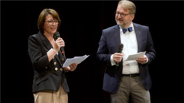 Eine Frau und ein Mann, beide mit Mikrofon und Redemanuskript in den Händen, auf einer Bühne.