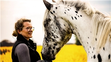 Eine Frau mit Brille steht lächelnd vor einem weißen Pferd mit schwarzen Punkten. Im Hintergrund ist ein gelbes Feld zu erkennen. 