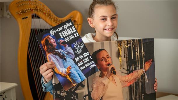 Dank des „Community-Tickets“ steht Georgia im Finale der Gesangsshow „The Voice Kids“. Damit möglichst viele anrufen, werden Plakate in der Stadt verteilt.
