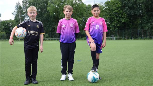 Drei Kinder stehen auf einem Fußballfeld. Einer von ihnen hält den Ball unter seinem Arm. Ein anderer steht mit einem Fuß auf dem Ball.  