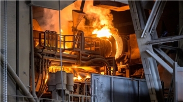 Blick ins Innere eines Stahlwerks: Glühende, orangefarbene Flammen und flüssiges Metall schieben sich durch Rohre und Walzen, umgeben von massiven Stahlträgern und Industrieanlagen.