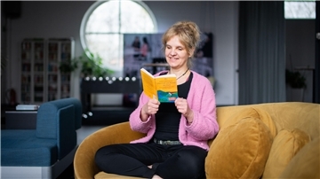 Eine Frau mit blonden Haaren sitzt auf einer gelben Couch und blickt in ein Buch. 