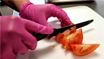 Ein Mensch schneidet mit einem Messer eine Tomate - dabei trägt er Einweg-Handschuhe.