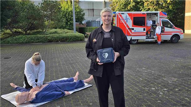 Eine Frau hält einen Defibrillator in den Händen, hinter ihr übt eine junge Frau an einer Reanimationspuppe die Herz-Druckmassage. Im Hintergrund steht ein Rettungswagen der Feuerwehr Bremerhaven mit zwei Einsatzkräften.