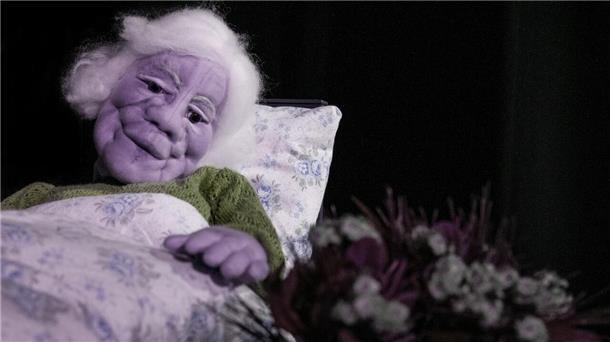 Zu sehen ist eine Handpuppe in Gestalt einer alten Frau, die im Bett liegt und auf einen Blumenstrauß blickt.