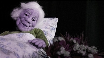 Zu sehen ist eine Handpuppe in Gestalt einer alten Frau, die im Bett liegt und auf einen Blumenstrauß blickt.