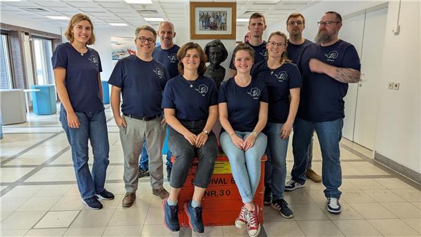 Eine Gruppe Menschen steht in einem Raum zusammen und lächelt in die Kamera. Sie tragen dunkelblaue T-Shirts. Es sind vier Frauen und fünf Männer.