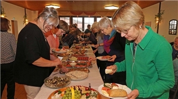 Eine Gruppe von Frauen steht an einem Buffet in einem Raum einer Gaststätte. Sie bedienen sich an einer großen Auswahl an Speisen, darunter Obst, Brot, Aufschnitt und verschiedene Salate, die auf einem langen Tisch präsentiert sind.