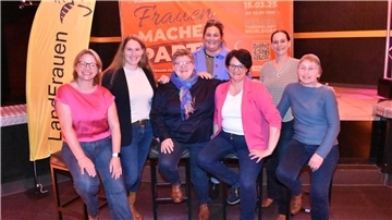 Sieben Frauen stehen und sitzen nebeneinander und lachen in die Kamera.