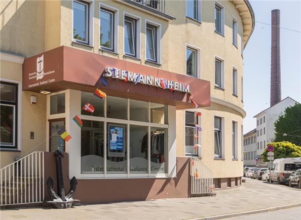 Ein gelbes Gebäude, auf dem steht: Seemannsheim. Im Hintergrund ist ein Schornstein zu sehen.