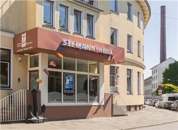 Ein gelbes Gebäude, auf dem steht: Seemannsheim. Im Hintergrund ist ein Schornstein zu sehen.