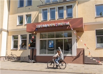 Ein gelbes Gebäude, an dem eine Frau auf einem Fahrrad vorbeifährt. Über dem Eingang steht "Seemannsheim".