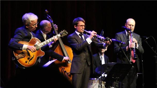 Vier Jazzmusiker auf einer Bühne: Ein Gitarrist, ein Kontrabassist, ein Trompeter und ein Klarinettist spielen gemeinsam in eleganter Abendkleidung, umgeben von Mikrofonen und Instrumenten.