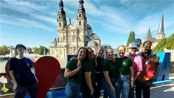Eine Gruppe junger Mitglieder des Jugendrotkreuzes posiert lachend mit ihrem Betreuer vor der barocken Fasanerie-Kulisse in Fulda, im Hintergrund die Kathedrale.