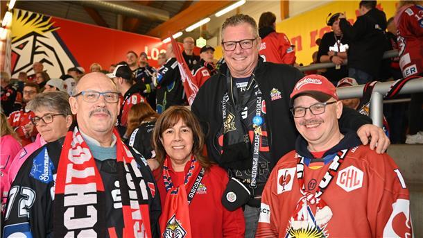 Drei Männer und eine Frau in Eishockey-Trikots stehen auf einer Tribüne.