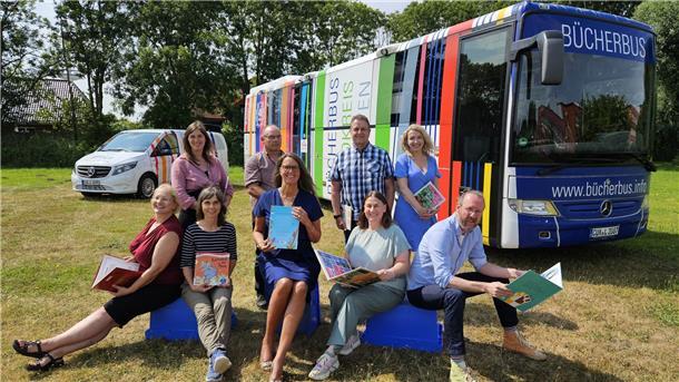 Bücherbus und Transporter „MobiLe“ stehen auf einer grünen Wiese. Davor sitzen und stehen neun Menschen, die jeweils Bücher in den Händen halten.