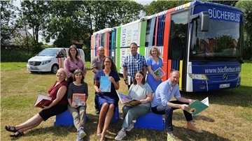 Bücherbus und Transporter „MobiLe“ stehen auf einer grünen Wiese. Davor sitzen und stehen neun Menschen, die jeweils Bücher in den Händen halten.