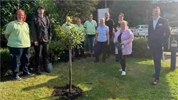 Mehrere Mitarbeitende und Bewohner der Pflegeeinrichtungen in Langen stehen bei Sonnenschein rund um einen frisch gepflanzten Apfelbaum auf einer Rasenfläche.