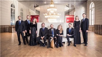 Neun Musiker stehen in schicker schwarzer Kleidung in einem festlichen Saal mit Kronleuchter nebeneinander. 