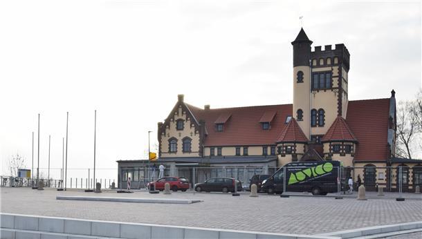 Ein Gebäude mit einem Turm steht an der Weser. Darum ein Bauzaun. Vor dem Gebäude stehen drei Autos.