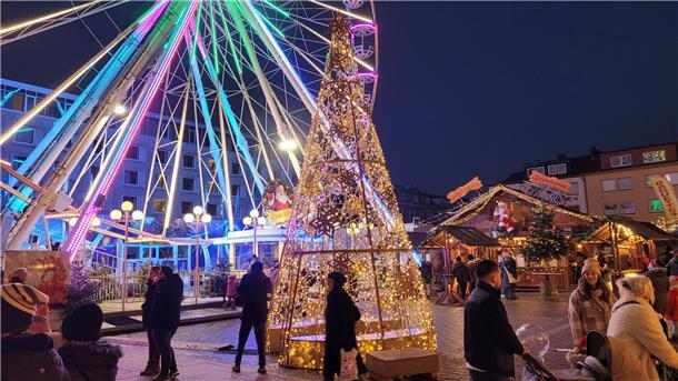 Riesenrad und Lichtkegel zwischen Buden auf dem Weihnachtsmarkt; dazwischen Menschen.