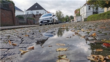 Kopfsteinpflaster mit großer Pfütze. Im Hintergrund: mehrere Autos am Straßenrand.