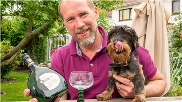 Ein Mann hält eine Flasche Wein in der rechten und seinen Dackel in der linken Hand.
