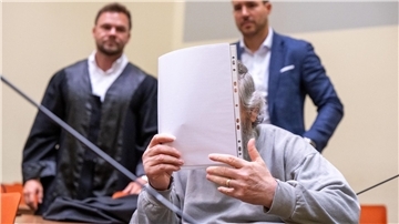 Der Angeklagte hält sich im Gerichtssaal ein Blatt Papier vor das Gesicht. Im Hintergrund stehen seine Anwälte.