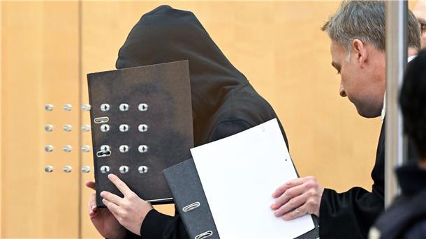 Der Angeklagte (l) verdeckt mit einem Aktenordner sein Gesicht, als er mit seinem Verteidiger zu Beginn der Verhandlung in einem Saal am Oberlandesgericht eintrifft.