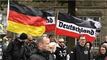 Teilnehmer einer rechtsextremen Demonstration mit schwarz-weiß-roter Flagge mit der Aufschrift „Deutschland“ und einer Deutschlandflagge; mehrere Personen in dunkler Kleidung, teils mit Sonnenbrillen oder Kapuzen, stehen eng beieinander.