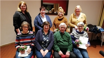 Eine Gruppe von acht Personen, überwiegend Frauen, posiert für ein Foto in einem Innenraum mit einer schlichten Wand und einem Spiegel im Hintergrund. Einige halten Blumensträuße und Urkunden in den Händen.