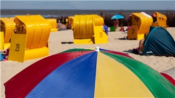 Ein bunter Sonnenschirm im Vordergrund, dahinter Sandstrand, gelbe Strandkörbe und Menschen, verschwommen.