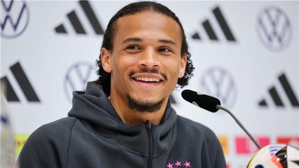 Nationalspieler Leroy Sané sitzt auf einem Podium und spricht mit Medienvertretern. 