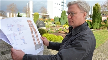 Der Elsdorfer Pastor Michael Grimm steht mit der Zeichnung des geplanten Glockenturms auf dem Friedhof. Der wird voraussichtlich Anfang kommenden Jahres gebaut. Im Hintergrund sind Gebäude der Molkerei zu sehen.