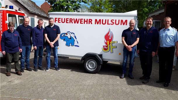 Mehrere Männer stehen vor einem Anhänger mit der Aufschrift „Feuerwehr Mulsum“ und den Logos der Jugend- und Kinderfeuerwehr; links ein Feuerwehrfahrzeug.