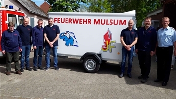 Mehrere Männer stehen vor einem Anhänger mit der Aufschrift „Feuerwehr Mulsum“ und den Logos der Jugend- und Kinderfeuerwehr; links ein Feuerwehrfahrzeug.