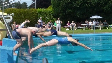 Mehrere Badegäste springen gleichzeitig ins Freibad Midlum, während Zuschauer im Hintergrund auf Stühlen das Geschehen verfolgen.