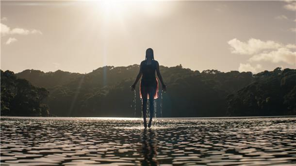 Eine Frau schwebt über einem See. Die Hände sind ein wenig ausgebreitet. Im Hintergrund ist ein Wald zu sehen. Die Sonne scheint. 