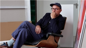 Ein Mann mit Brille, dunkler Jacke und Mütze sitzt nachdenklich in einem Designerstuhl. Er hat ein Bein über das andere geschlagen und stützt sein Kinn auf die Hand. Im Hintergrund sind Leinwände und ein Kunstwerk zu sehen.