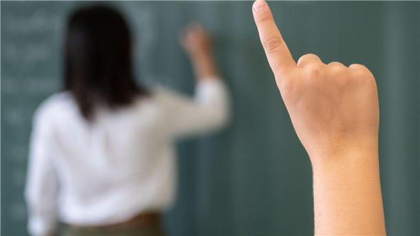 Ein Schüler meldet sich, während die Lehrerin an die Tafel schreibt.
