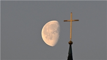 Der Mond im Morgenlicht hinter einem Kreuz auf einem Kirchturm.