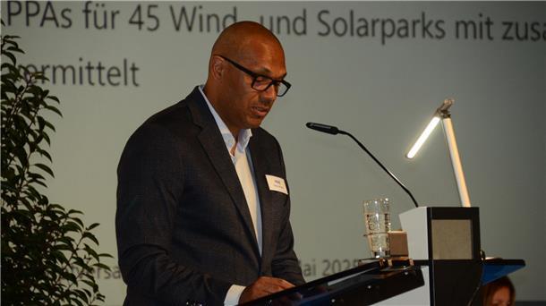 **ALT-Text:**
Ein Redner spricht bei einer Präsentation am Mikrofon, vor sich ein Glas Wasser und ein Rednerpult, im Hintergrund ist ein Text zur Energiewirtschaft projiziert.