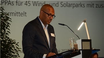 **ALT-Text:**
Ein Redner spricht bei einer Präsentation am Mikrofon, vor sich ein Glas Wasser und ein Rednerpult, im Hintergrund ist ein Text zur Energiewirtschaft projiziert.