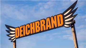 Ein Schild zwischen zwei Holzpfählen mit der Aufschrift "Deichbrand". An der Seite ist das Schild künstlerisch mit Flügeln verziert. Im Hintergrund blauer Himmel mit leichten Wolken.